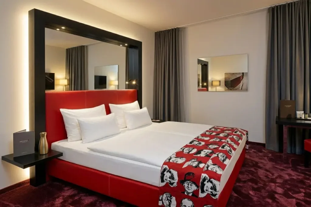 A modern hotel room at Arcotel Nike Linz featuring a vibrant red leather headboard, a large mirror frame with built-in lighting, and dark grey curtains.