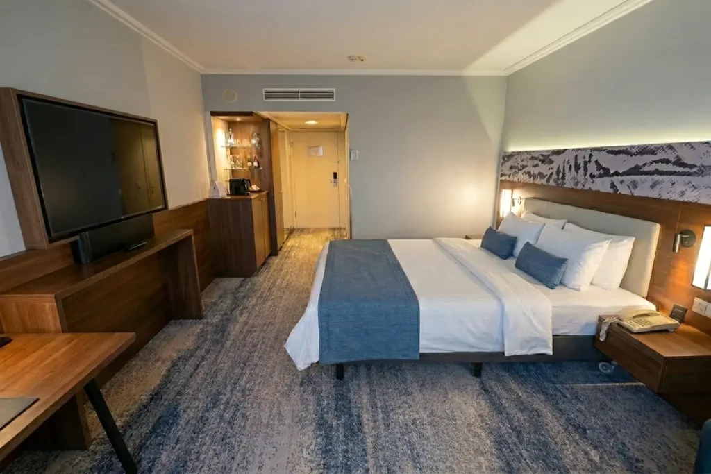 modern, premium hotel room at Courtyard by Marriott Linz featuring a king bed with blue accent pillows, textured blue carpeting, dark wood paneling, and a large flat-screen TV.