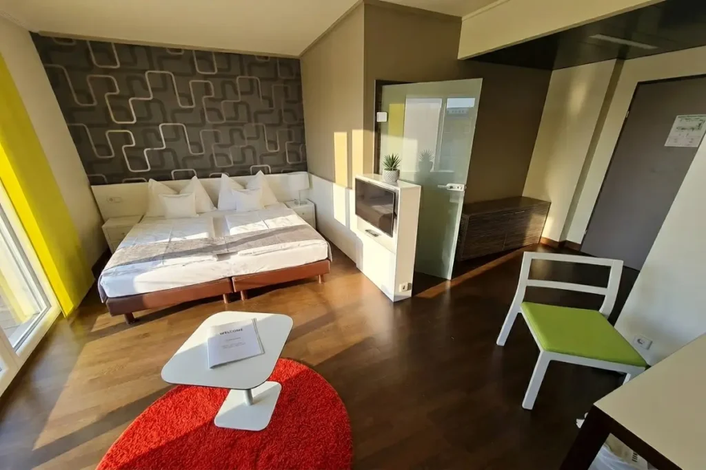 A modern, sunlit studio hotel room at Harry's Home Linz-Urfahr featuring a large bed, patterned wallpaper, a red circular rug, and a green accent chair.