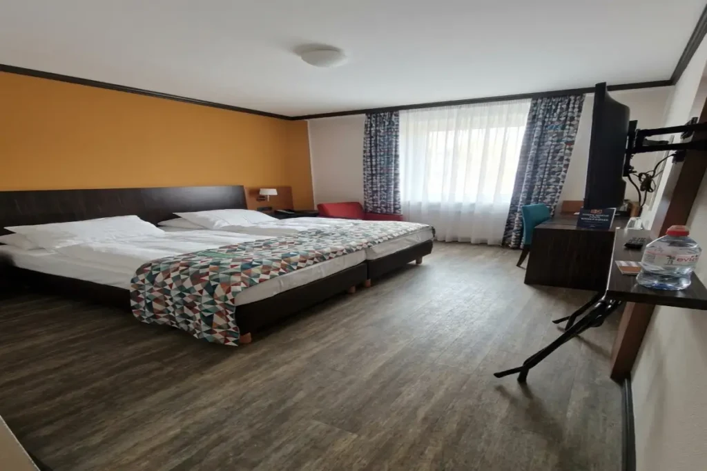 A comfortable hotel room at Trans World Hotel Donauwelle featuring a geometric patterned bed runner, light wood flooring, and an orange accent wall.
