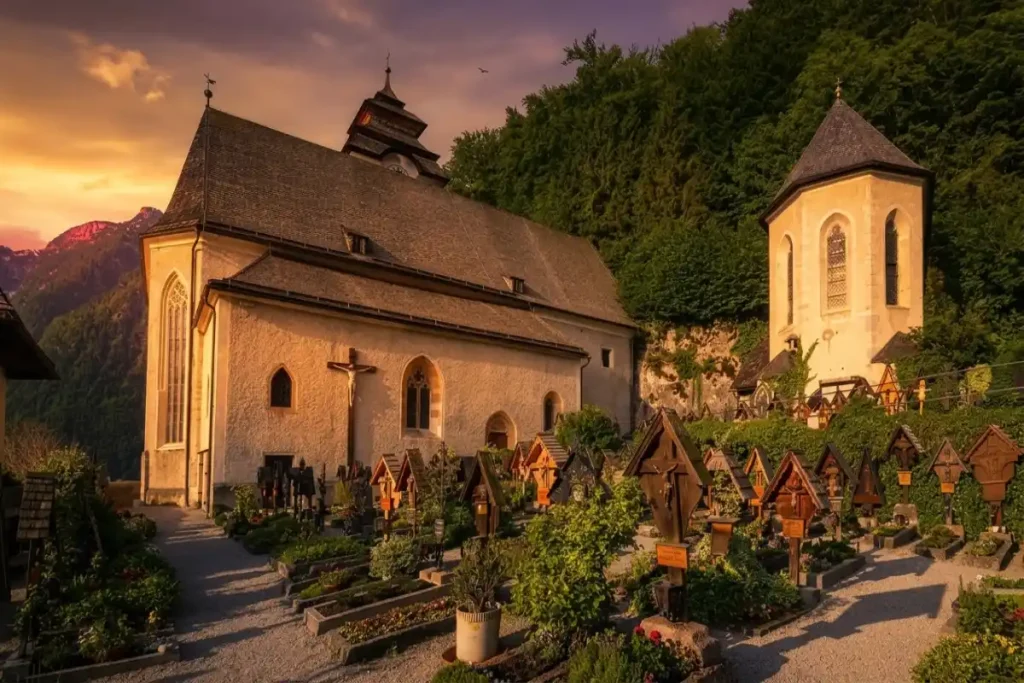 Catholic Church and flower-filled historic cemetery in Hallstatt, surrounded by forested alpine mountains at sunset.